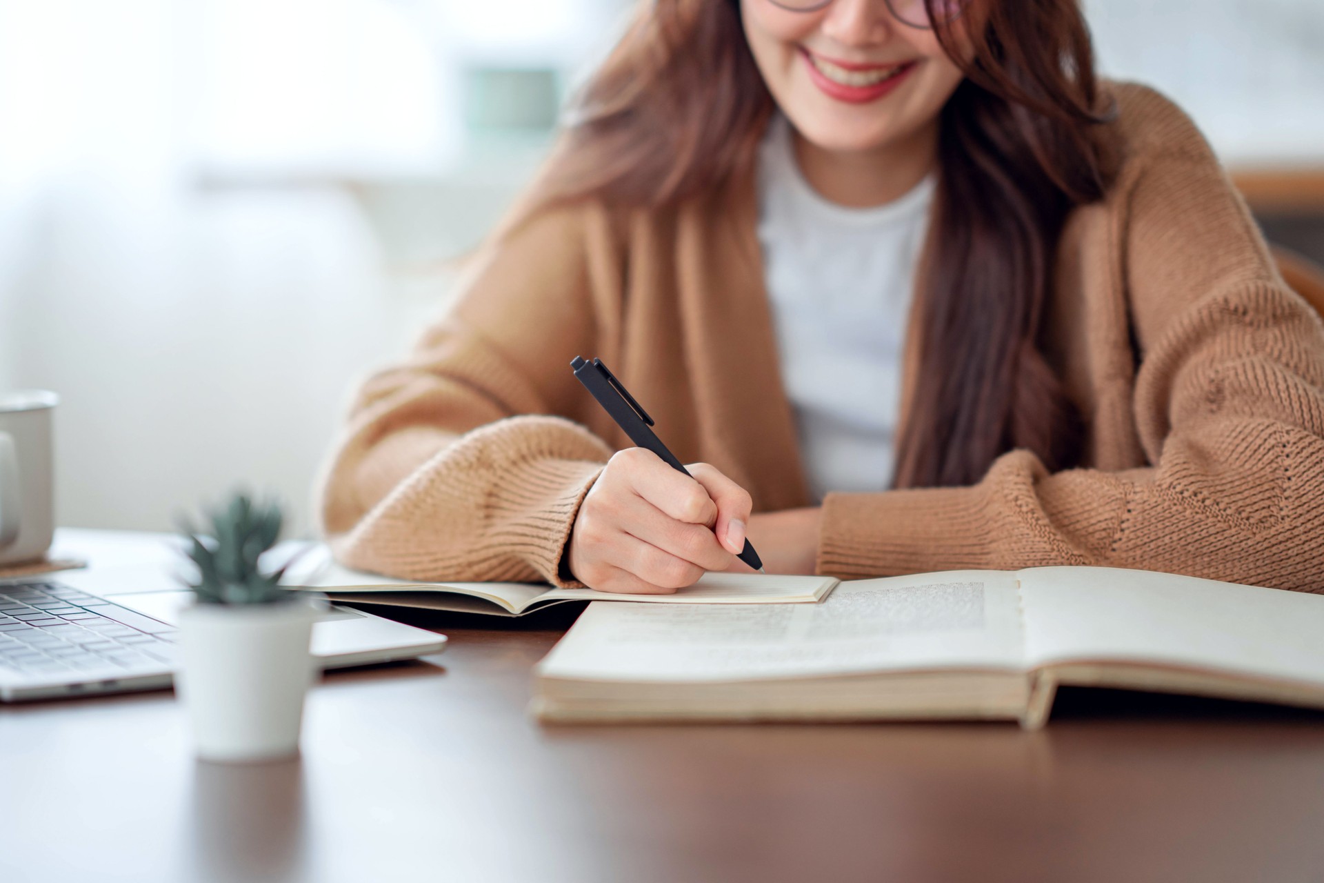 Hand of woman writing in spiral notepad placed on wooden desktop with various items at home.